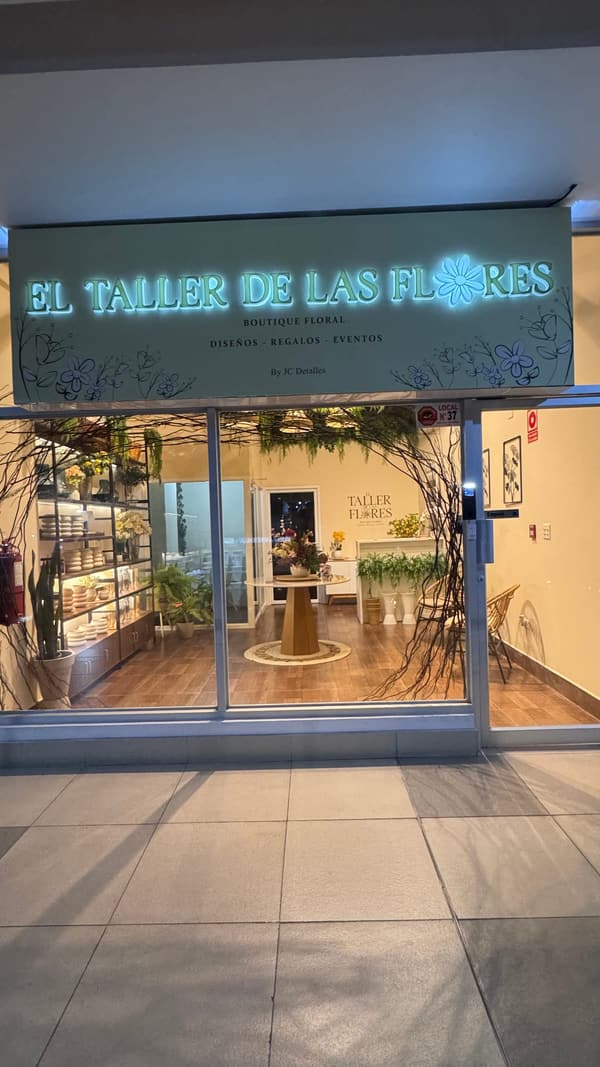 Fachada iluminada de El Taller de las Flores en Plaza Lefevre: el rótulo de la boutique y, tras la vitrina, la mesa con un arreglo y los estantes de floreros